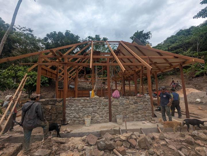 Las cerchas del techo puestas y el muro de piedra empezando, con el equipo (y los perros) trabajando.